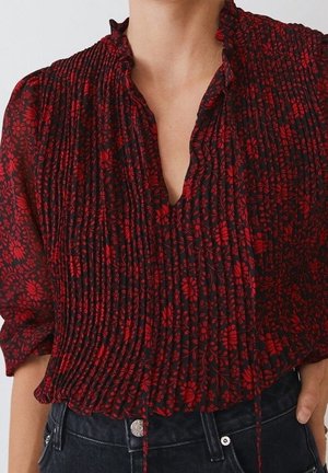 Femme portant un chemisier plissé rouge à fleurs avec un col en V et un col à volants, associé à un jean foncé taille haute avec boutons visibles.