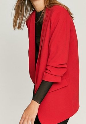 Persoon draagt een felrode blazer over een zwarte top met lange mouwen, met de mouwen van de blazer nonchalant opgerold.