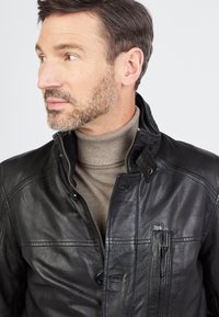 Schwarze Lederjacke mit Stehkragen, Reißverschluss und vorderen Taschendetails. Getragen über einem beigen Rollkragenpullover.