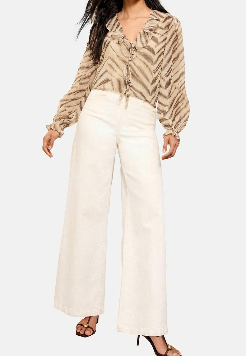 Blusa beige a righe con colletto arricciato e maniche lunghe e voluminose, abbinata a pantaloni bianchi a gamba larga e sandali neri con cinturini.