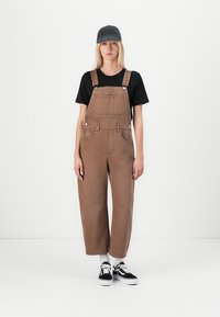 Bruine denim tuinbroeken met een losse fit, verstelbare banden en voorkeurszakken, gecombineerd met een zwart t-shirt en een grijze pet, en zwarte sneakers.