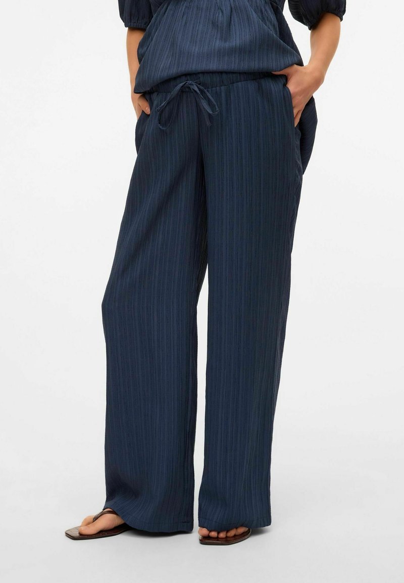 Pantalon large bleu marine à cordon avec texture verticale, porté les mains dans les poches et sandales marron à bout ouvert.
