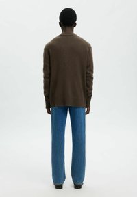 Persona che indossa un maglione marrone a maniche lunghe, jeans blu dritti e scarpe nere, in piedi girato di spalle su uno sfondo semplice.
