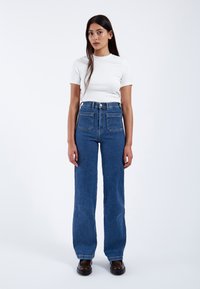 Vaqueros de mezclilla azul de tiro alto y pierna recta, con cuatro bolsillos, costuras detalladas y un acabado ligeramente desteñido. Combinados con una camiseta blanca ajustada.