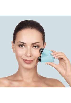 Femme à la peau claire utilise un masseur facial bleu avec deux rouleaux noirs sur la joue, sur fond bleu clair.