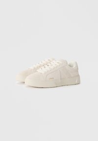 Sneaker low-top in suede bianco con punta arrotondata, lacci piatti e suola in gomma texture. Dettaglio del logo oro sul lato.