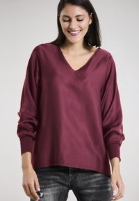 Burgundy V-neck top made of smooth fabric, with long, slightly puffed sleeves and elastic cuffs, paired with dark denim jeans.