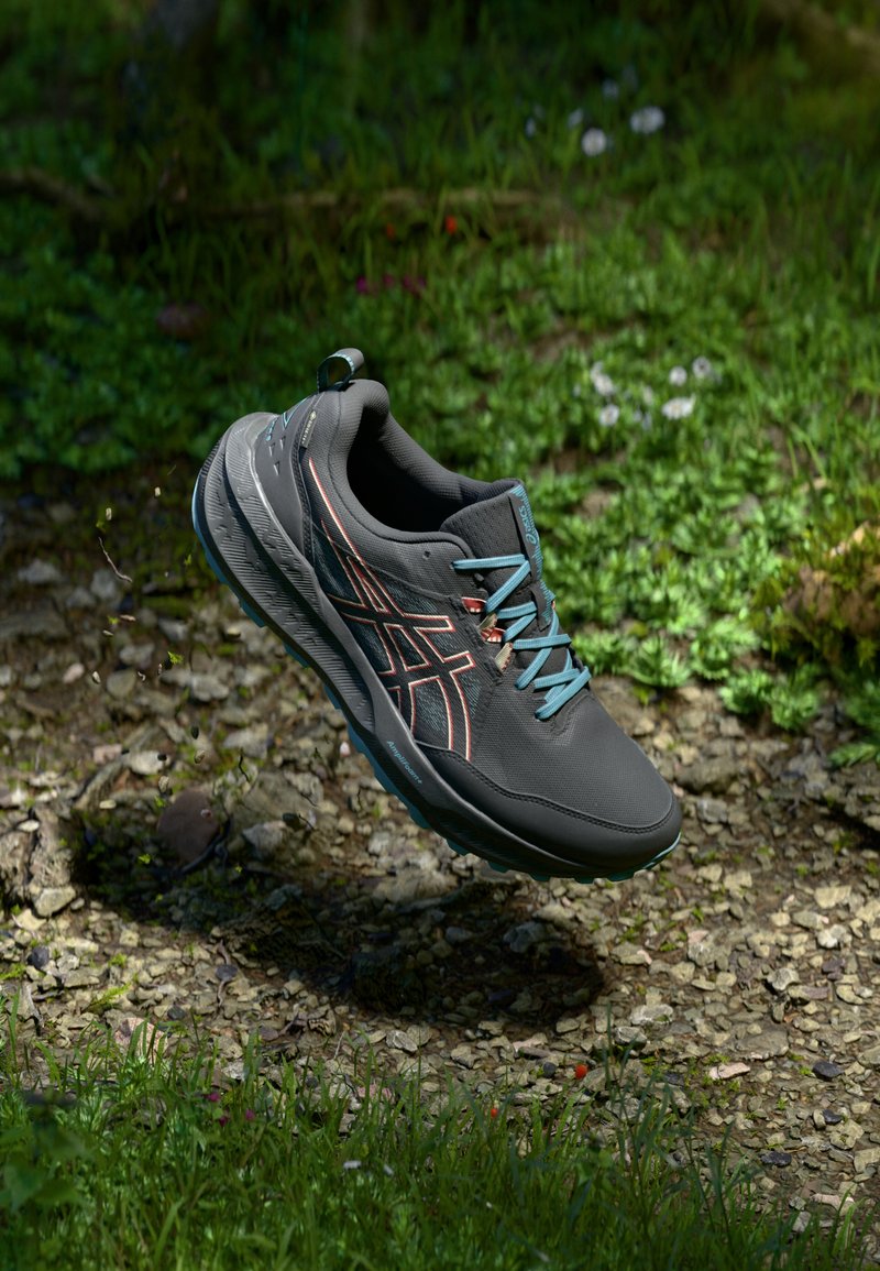 Zapatilla deportiva negra con cordones turquesas, suela gris texturizada y detalles en naranja, suspendida sobre una superficie natural con vegetación.