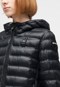 Νεαρό άτομο με κοντά κόκκινα μαλλιά που φοράει μαύρο επενδεδυμένο μπουφάν με κουκούλα και λογότυπα Blauer USA στο στήθος και στο μανίκι.