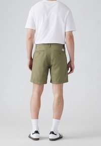 Oliivinvihreät shortsit suoran leikkauksen kanssa, joissa on kaksi takataskua ja merkkilogo. Yhdistetty valkoisen t-paidan ja valkoisten sukkien kanssa.