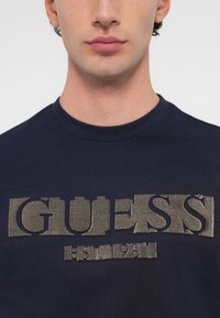 Tumed sinine puuvillane T-särk, mille esiküljel on tekstureeritud beež "GUESS" logo ja asutamisaasta "1981", ümar kaelus.