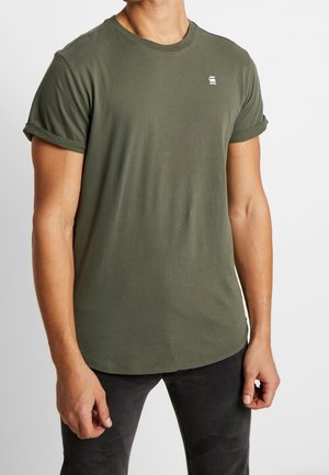 Homme portant un t-shirt vert olive à manches courtes avec des revers et un petit logo blanc sur la poitrine, associé à un pantalon noir.