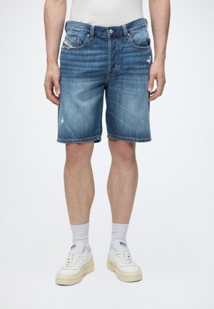 Homme portant un short en jean bleu jusqu'aux genoux, des chaussettes blanches et des baskets blanches, debout sur un fond uni.