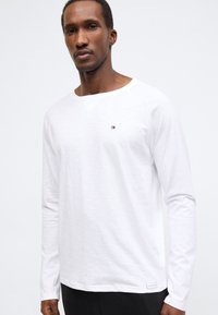 Λευκή μακρυμάνικη μπλούζα με στρογγυλή λαιμόκοψη, με υφή υφάσματος και μικρό λογότυπο στο στήθος. Κανονική εφαρμογή, με ίσια τελείωμα.