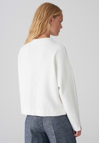 White, oversized sweater with a high neckline and dropped shoulders; smooth texture, ribbed cuffs, and a central seam at the back.