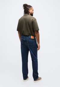 Άνδρας με κοτσίδες φοράει πόλο πουκάμισο χακί, σκούρο μπλε τζιν και μπεζ αθλητικά παπούτσια, στέκεται και κοιτάει προς τα πλάγια σε λευκό φόντο.