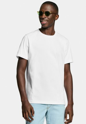 Young man wearing sunglasses, a plain white t-shirt, and light blue jeans, smiling and looking to the side against a white background.