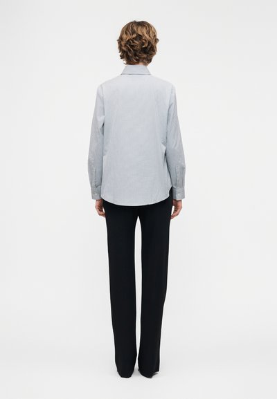 Άτομο με κοντά σγουρά μαλλιά, που φοράει ανοιχτό μπλε πουκάμισο με μακριά μανίκια και μαύρο παντελόνι ίσιας γραμμής, στέκεται με την πλάτη προς το μέρος μας σε λευκό φόντο.