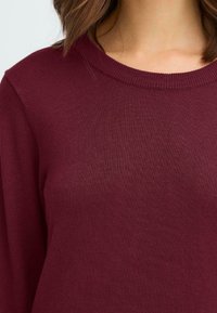 Bordeaux gebreide trui met een ronde hals en lange mouwen, met een zachte textuur en ribbelstructuur bij de kraag.