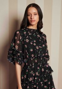 Ung kvinna med långt mörkt hår klädd i en svart klänning med rosa blommönster och volangdetaljer, stående mot en beige randig vägg.