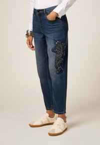 Vrouw in donkerblauwe jeans met donkere bloemenborduursels op de dij, witte shirt, armbanden en witte sneakers met gumzolen.