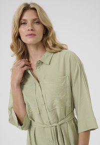 Abito camicia verde chiaro con colletto, chiusura a bottoni, tasca e vita regolabile con cintura. Il tessuto ha una finitura texturizzata e maniche a 3/4.