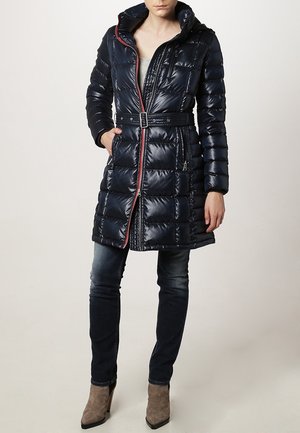 Femme portant un manteau matelassé bleu marine brillant ceinturé avec une capuche, associé à un jean skinny foncé et des bottines taupe à talons.