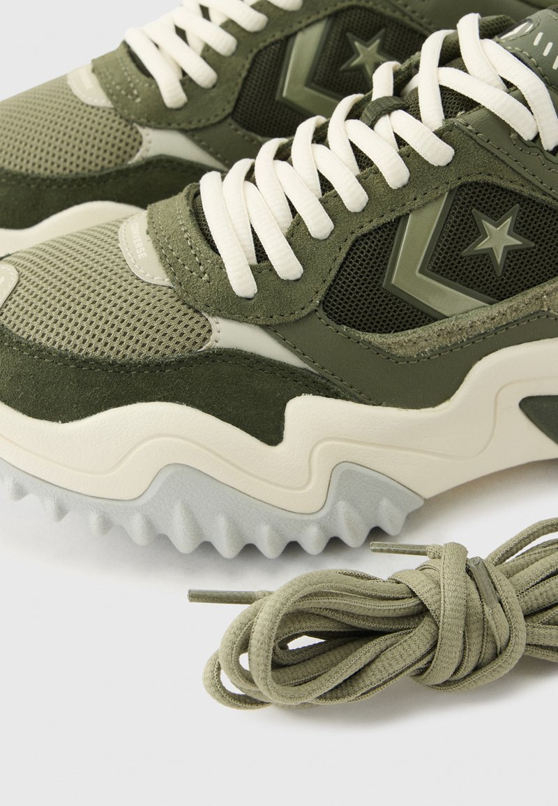 Olivgrüne und beige Sneakers mit weißen Schnürsenkeln, Sternlogo an der Seite, strukturierter Sohle und zusätzlich olivgrünen Schnürsenkeln neben ihnen.