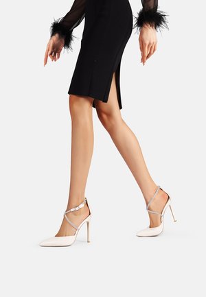 Scarpe con tacco alto in vernice bianca, con punta affilata, sottile tacco a stiletto e una cinghia alla caviglia regolabile con accento metallico.