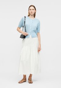 Femme portant un cardigan à manches courtes bleu clair boutonné, une longue jupe blanche, des sandales marron à lanières, et portant un petit sac à bandoulière noir.