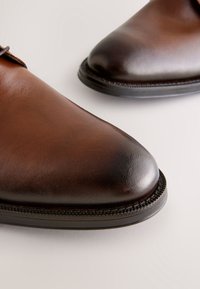 Brune lædersko med en poleret finish, spids tå og detaljerede syninger langs kanten af sålen. Glat tekstur overalt.