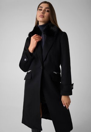 Mujer con un abrigo largo negro con cuello de piel negra, bolsillos con cremallera y puños con botones, posando frente a un fondo gris liso.