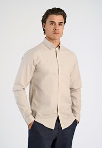 Beige Langarmhemd mit Knopfkragen, aus einem strukturierten Stoff gefertigt. Verfügt über Vorderknöpfe und einen geraden Saum, getragen mit dunklen Hosen.