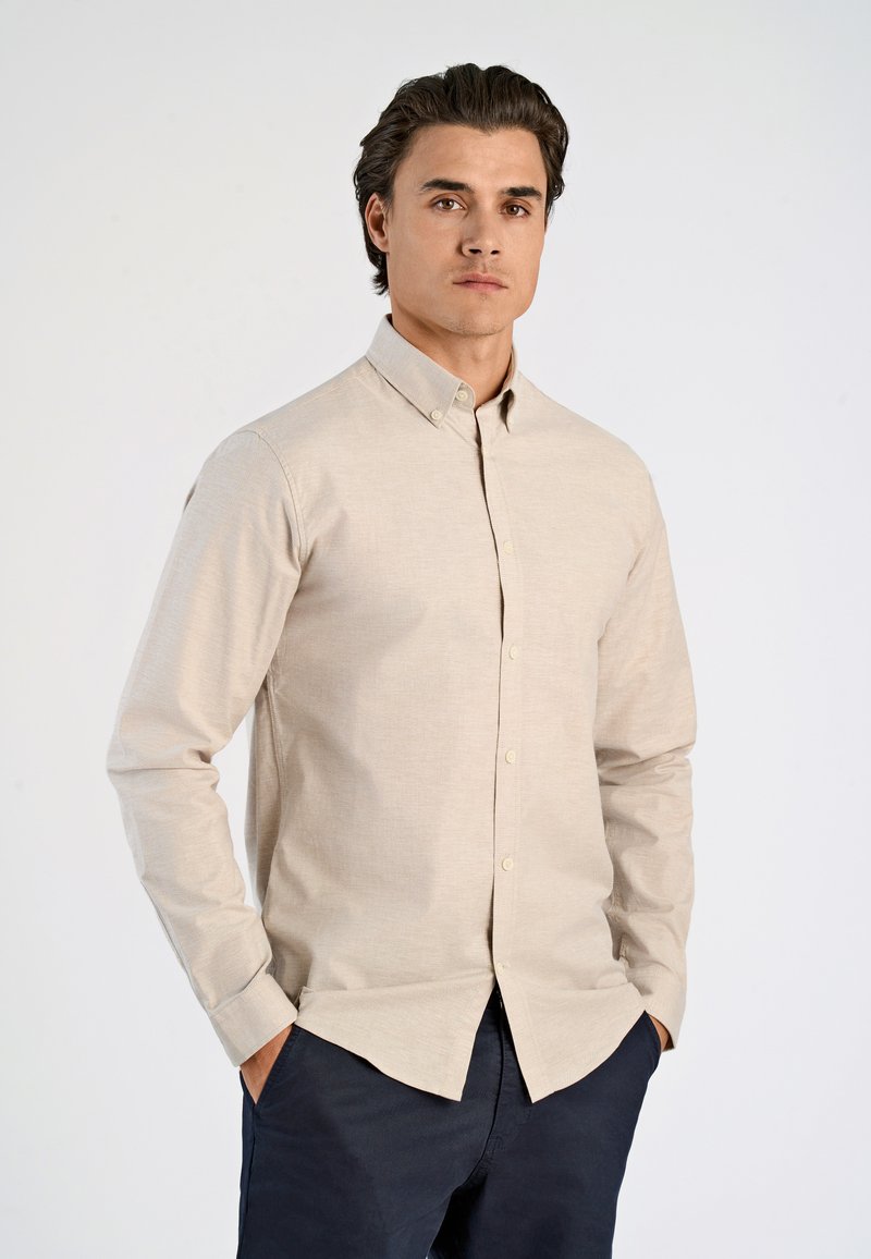 Beige Langarmhemd mit Knopfkragen, aus einem strukturierten Stoff gefertigt. Verfügt über Vorderknöpfe und einen geraden Saum, getragen mit dunklen Hosen.