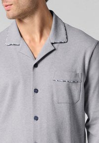Camicia grigia chiaro a maniche corte con bottone frontale, bottoni blu navy e taschino sul petto con bordo decorativo lungo i lati.