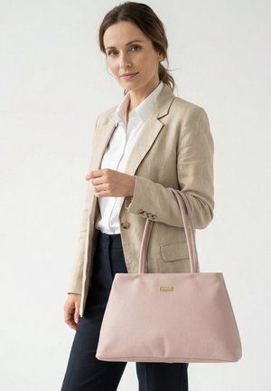 Vrouw met beige blazer en wit overhemd houdt een grote lichtroze handtas met dubbele handvatten vast tegen een egale achtergrond.