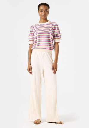 Pull en tricot rayé rouge, bleu et blanc, associé à un pantalon large plissé couleur crème. Modèle se tenant sur des sandales plates, fond neutre.
