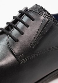 Zapato negro de cuero con tela negra texturizada cerca de los cordones, punta redonda, superficie lisa y seis cordones negros. Presenta costuras en contraste.