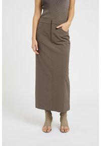 Jupe marron mi-longue en tissu lisse avec une taille ajustée et des poches avant. Associée à des chaussures ouvertes beiges.