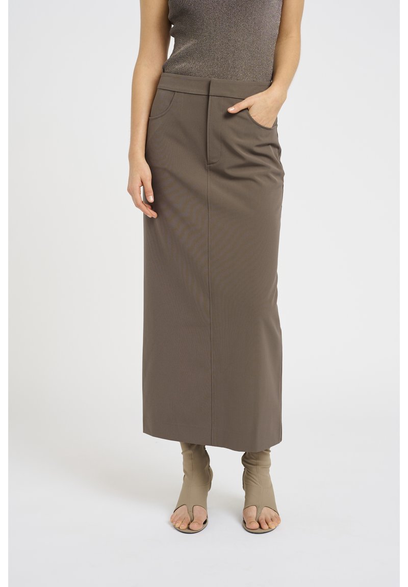 Jupe marron mi-longue en tissu lisse avec une taille ajustée et des poches avant. Associée à des chaussures ouvertes beiges.