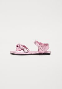 Rosa metallisk sandal med en knuten detalj fram, öppna sidor, justerbart band och en platt svart sula. Slät textur.