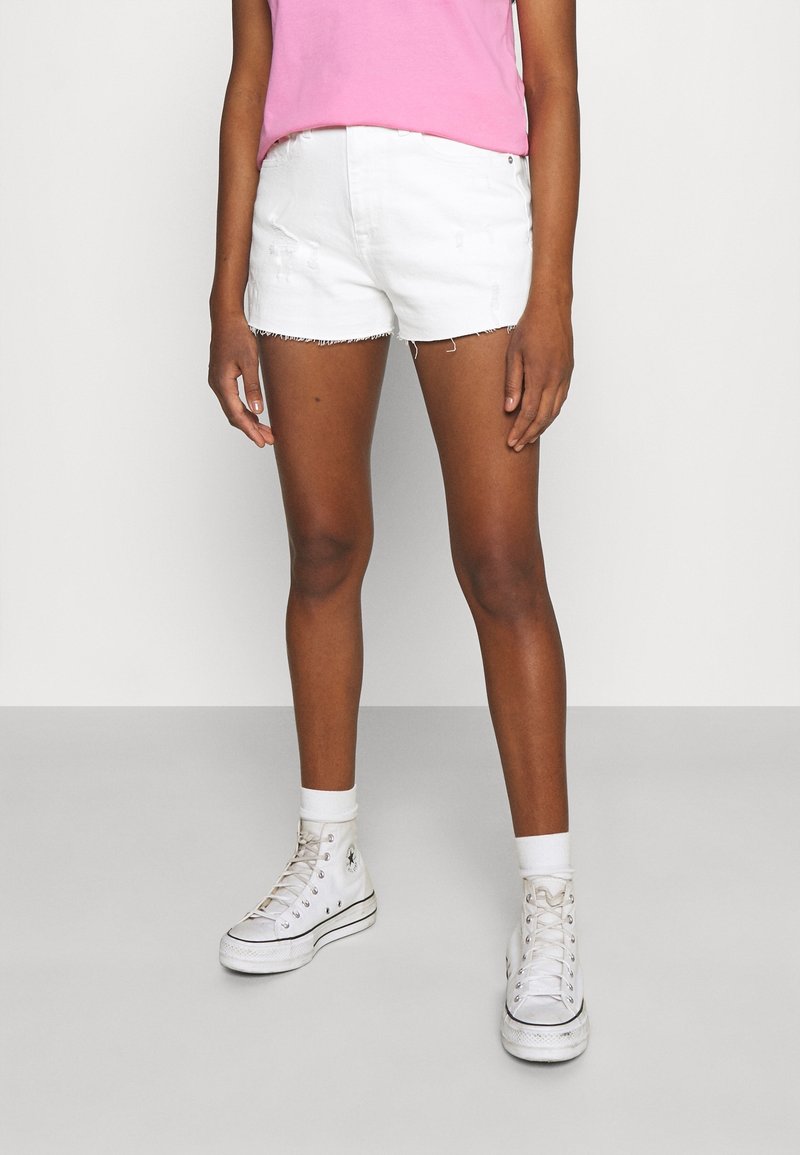 Personne portant un short en jean blanc, des baskets hautes blanches avec des chaussettes blanches, et un haut rose, debout devant un fond uni.
