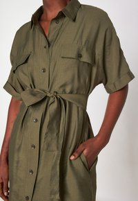 Abito camicia verde oliva con maniche corte, chiusura a bottoni frontale, due tasche sul petto, dettaglio in vita legato e tessuto morbido e liscio.