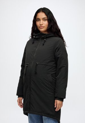 Mujer con cabello largo y oscuro que lleva un abrigo de invierno negro con capucha, cremallera frontal y cordones, de pie contra un fondo liso y claro.