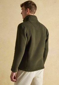 Tmavozelený fleecový pulóver s vysokým golierom a jemne embosovaným logom na zadnej strane. Má rovný lem a priliehavé rukávy.