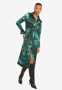 Rochie cămașă din satin verde, cu talie ajustabilă prin cordon și închidere cu nasturi. Prezintă un imprimeu animal vibrant și o crăpătură înaltă până la coapsă. Asortată cu cizme negre până la genunchi.