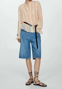 Chemisier long à manches longues de couleur pêche à motif floral avec ourlet à volants, associé à un short en denim bleu et des sandales noires à brides. Une ceinture noire souligne la taille.