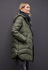Parka verde acolchado con capucha, con diseño acolchado, bolsillos laterales y puños de canalé. Llevada sobre una bufanda oscura frente a un fondo gris.