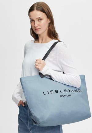 Giovane donna che indossa una maglia bianca a maniche lunghe e tiene una grande borsa a spalla Liebeskind Berlin azzurro chiaro con manici neri.