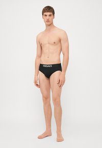 Svarte badetruser i stoff med en hvit "VERSACE"-logo på midjebåndet, glatt tekstur, tettsittende design, minimal dekning.